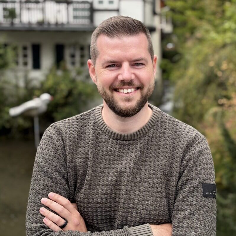 Mann mit kurzen Haaren und Bart trägt grauen, strukturierten Pullover mit rundem Kragen und schwarzem Ring an linker Hand, Arme verschränkt.