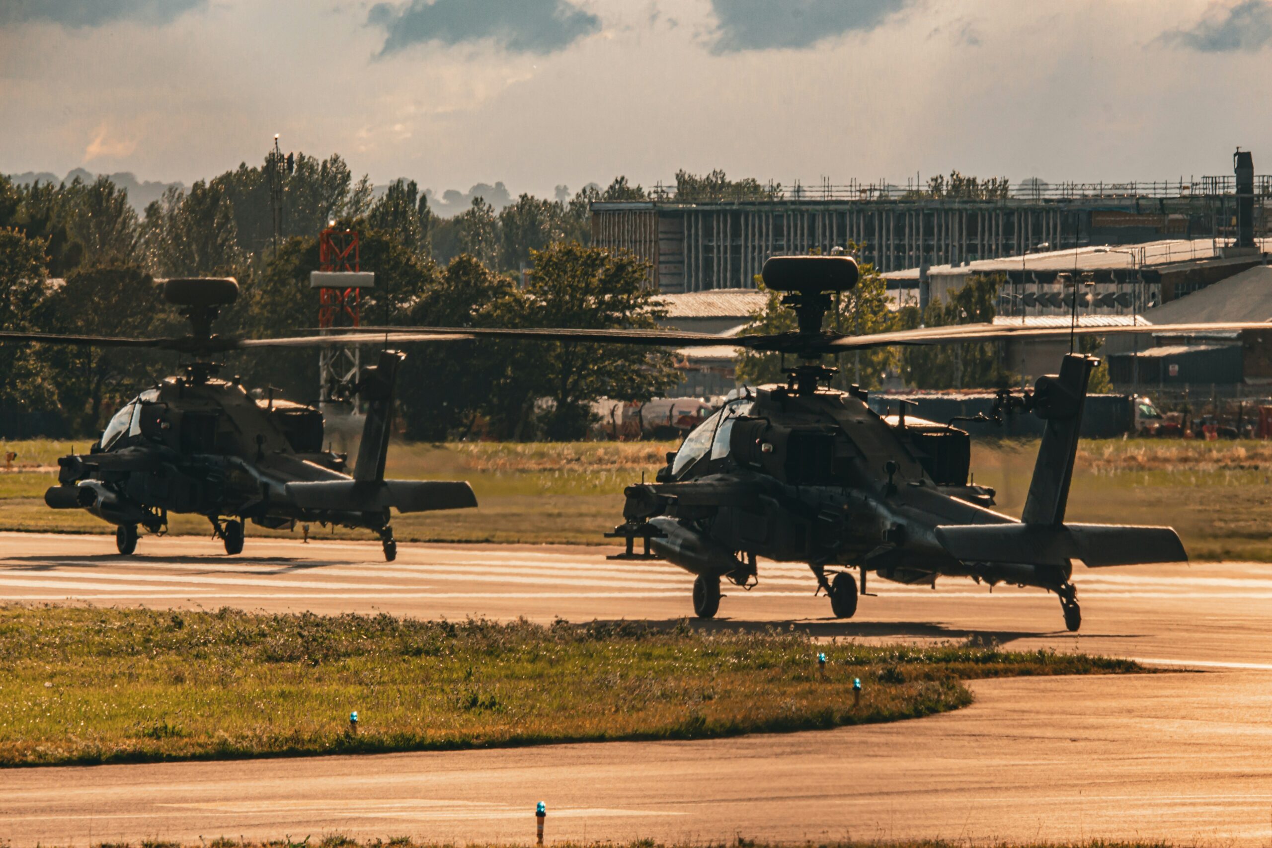 Zwei schwarze Kampfhubschrauber auf Startbahn vor Industriegebäuden und Bäumen bei bewölktem Himmel.